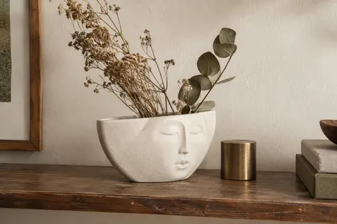 Petit vase blanc en céramique au visage féminin posé sur une étagère en bois patiné avec quelques tiges séchées.