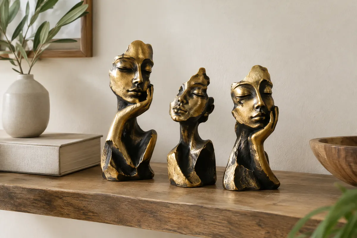 Trois sculptures abstraites de visages pensifs, en résine dorée et noire, posées sur une étagère en bois.