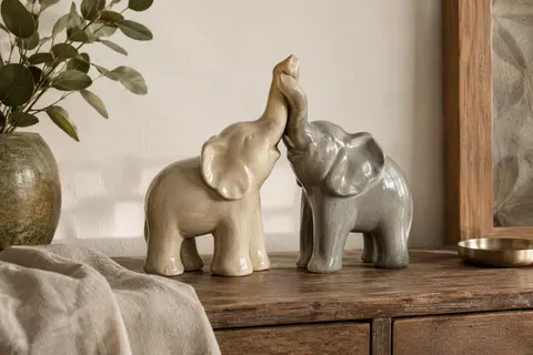 Deux éléphants en céramique beige brillante et gris doux, aux trompes enlacées, posés sur une console.