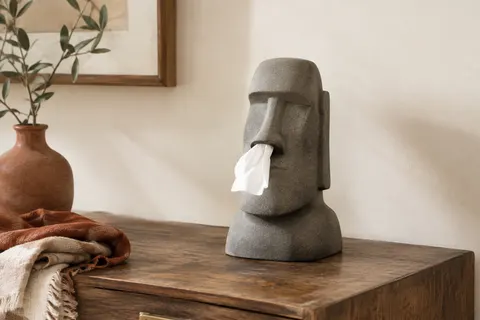 Distributeur de mouchoirs gris en forme de tête Moai posé sur une console en bois.