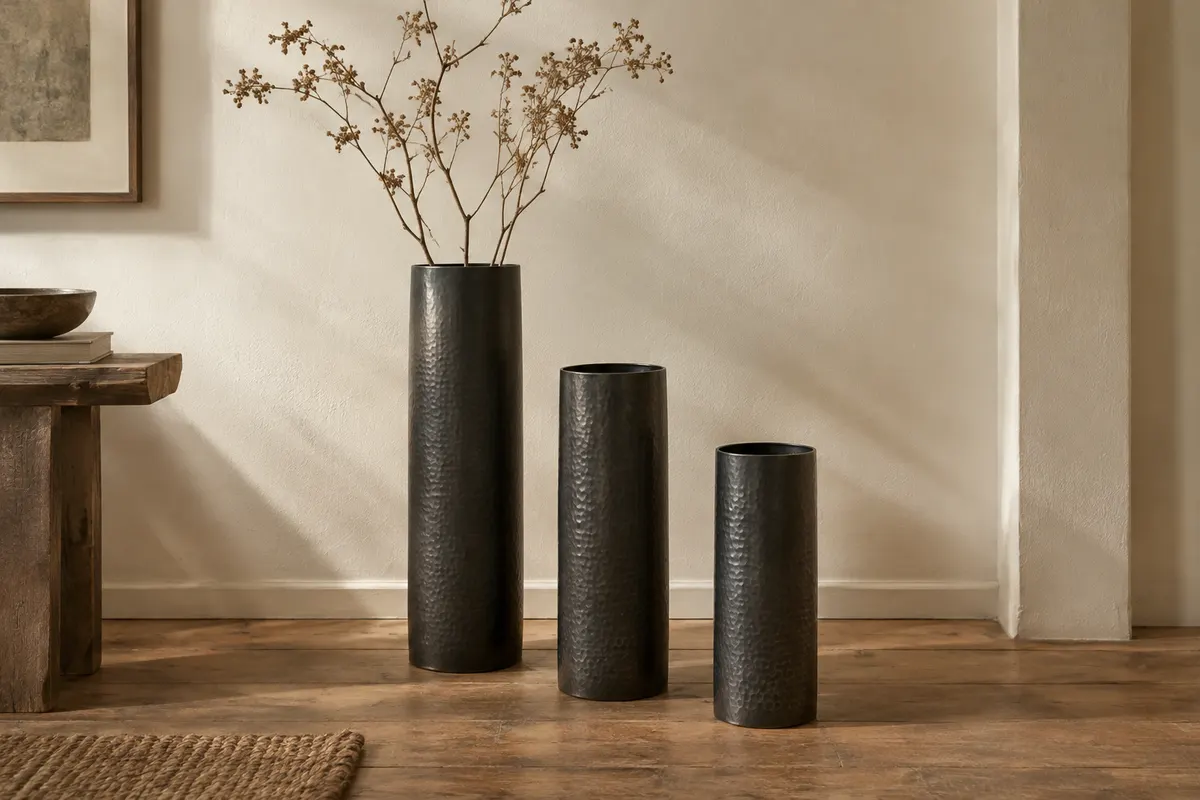 Trois vases cylindriques en métal noir texturé, de hauteurs différentes, posés près d’un mur ivoire.