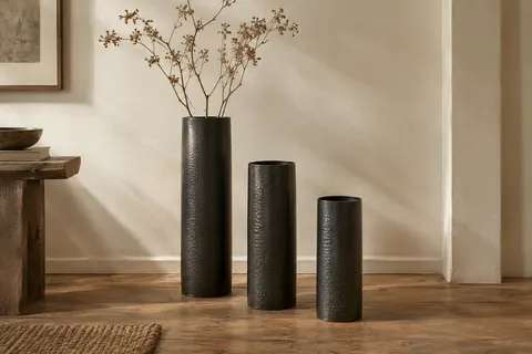 Trois vases cylindriques en métal noir texturé, de hauteurs différentes, posés près d’un mur ivoire.