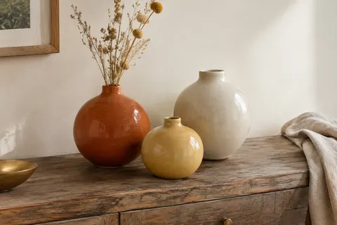 Trois vases ronds en céramique glacée, orange, jaune et crème, posés sur une console en bois.