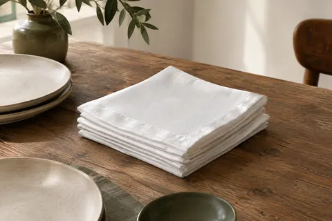 Serviettes de table carrées en coton blanc avec bord satiné, pliées sur une table en bois patiné.