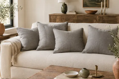 Quatre housses carrées en lin gris disposées sur un canapé clair dans un intérieur lumineux.