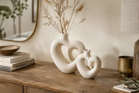 Duo de vases blancs en céramique mate aux formes de cœur imbriquées sur une console en bois.
